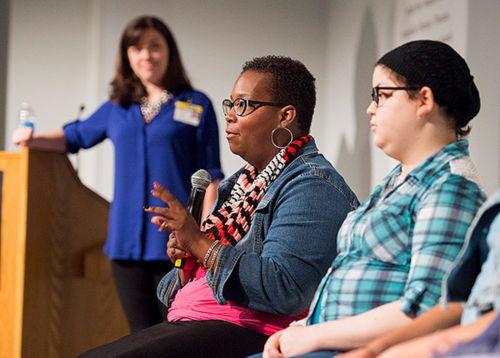 Photo of two young women sitting on a stage as a third person stands by a podium. 