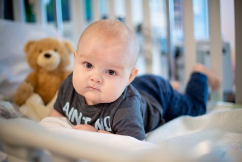 Male infant in crib in hospital setting with stuffed teddy bear in background