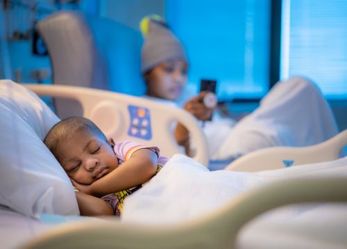 Little african american boy asleep in hospital bed with relative out of focus in background