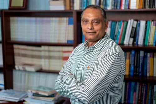 Deo Kumar Srivastava standing in front of a bookcase with his arms folded, looking at the camera and wearing a striped green shirt.