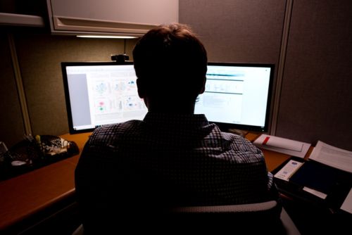Biostatistician sitting at the computer, back to the camera, two monitors illuminated in low light