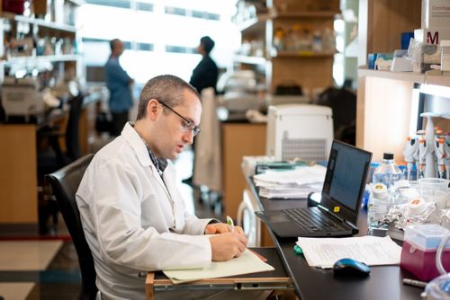 A male scientist sits at the bench in front of his laptop, writing notes in his notebook.