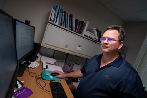 A man wearing glasses standing in front of two monitors, looking at biostatistical work
