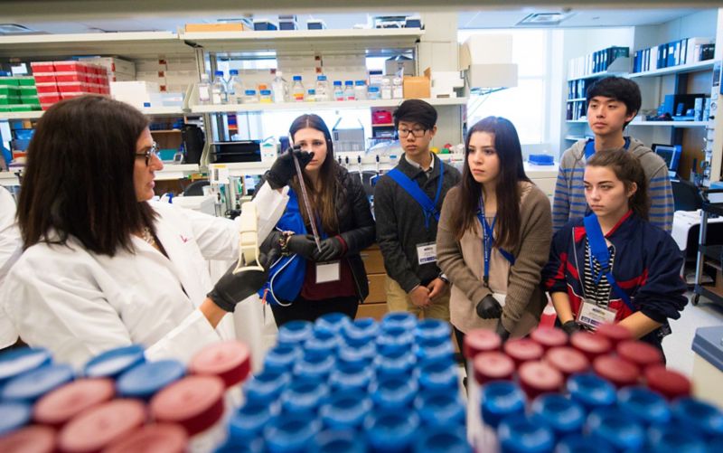 High school students listen to instructor in lab next to table full of test tubes.