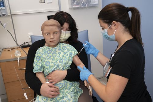 Mother holds a child dummy doll in her lap while nurse pretends to give the doll a shot