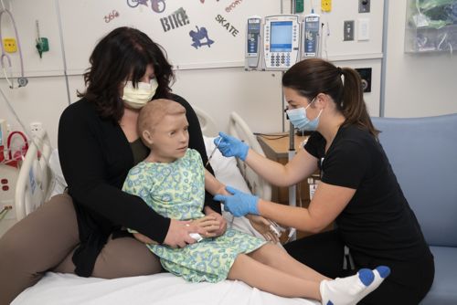 Mother holds child dummy doll in front of her on hospital bed while nurse pretends to give doll a shot