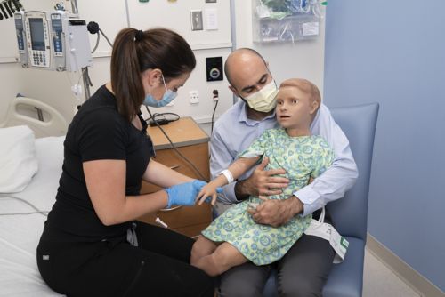 Man holds child dummy doll in lap with legs off to the side while nurse pretends to give a shot.