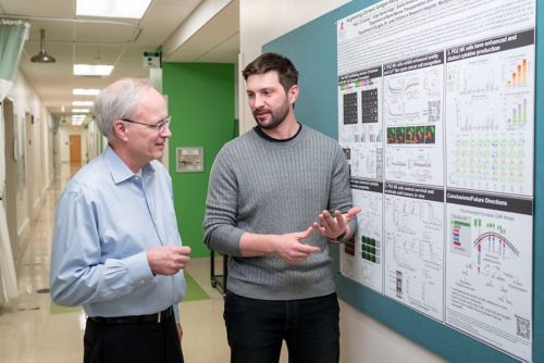 A man in a blue shirt talks to man in a gray shirt next to a bulletin board