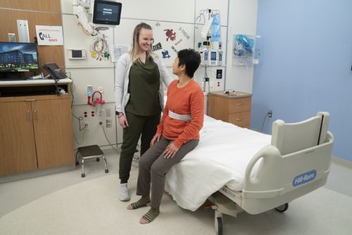 Woman, seated on bed, speaks to nurse who is standing.