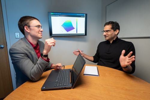 Two men sitting at a table with a computer having a conversation