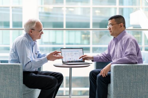 A man in a purple shirt discusses material with a man in a blue shirt with a computer in between them