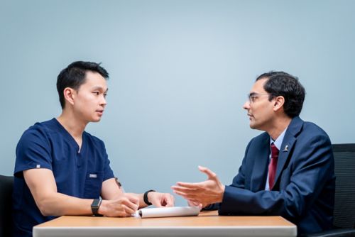 two men talking at a table. One is wearing hospital scrubs and is taking notes while the other, in a suit, talks. 