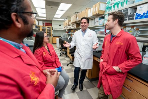 Group of people in a lab, some wearing red lab coats, Victor Torres in middle wearing a white one