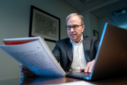 A man in a gray suit reads a notebook with his hand on a computer