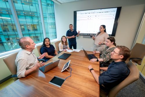 Meeting in conference type room. People around table, 2 presenting at screen.