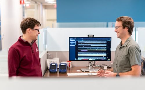 two men talking in front of a double screen computer monitor