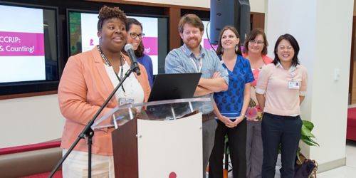 Photo of a group of people standing by a podium while a woman speaks