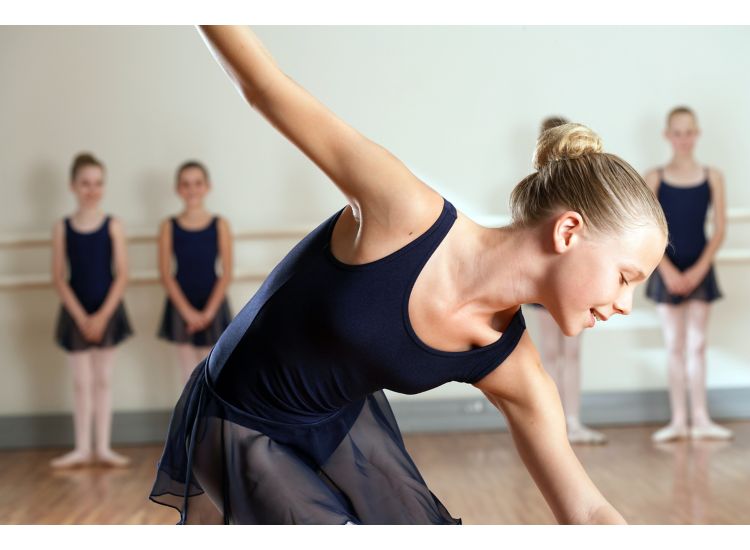 Young ballerina in foreground poses, four ballerinas in background stand against ballet bar