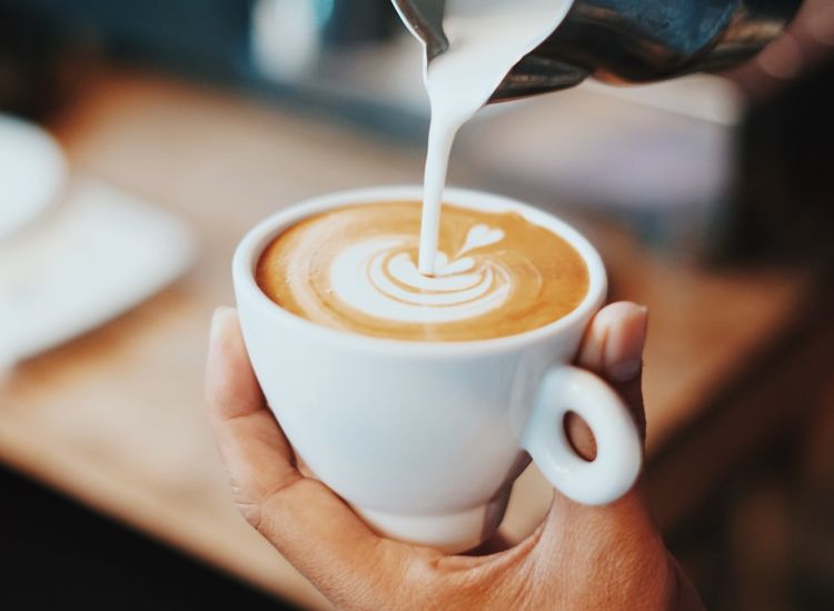 Hand holding white cup of coffee and pouring milk into a design