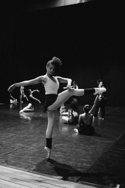 Diverse group of ballet dancers on a stage preparing to practice. Black dancer in point shoes in the forground posing on one leg. 