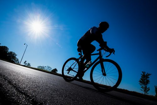 This picture shows a cyclist on a road bike wearing a helmet under a blue sky with a bright sun.