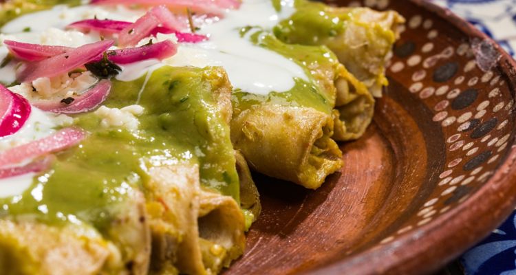 Close up of plated taquitos with green chile sauce, crema and pickled red onions.