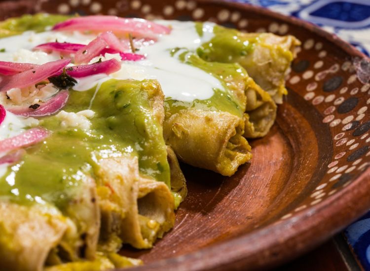 Close up of plated taquitos with green chile sauce, crema and pickled red onions.