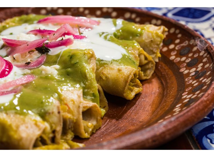 Close up of plated taquitos with green chile sauce, crema and pickled red onions.