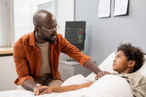 Young father supporting son who is lying in hospital bed