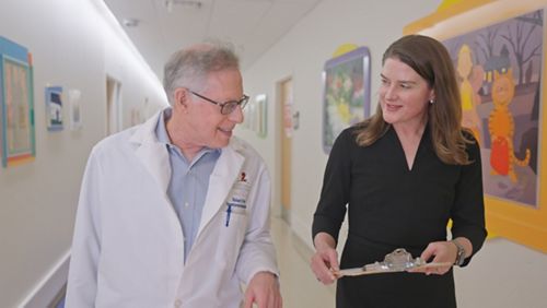 A man and woman in a hall way. The man is wearing a white coat. The woman is holding a clipboard