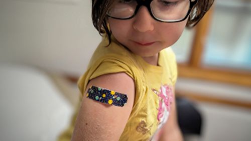 Young child wearing glasses and yellow shirt showing colorful bandage on upper arm after vaccination, pediatric immunization visit