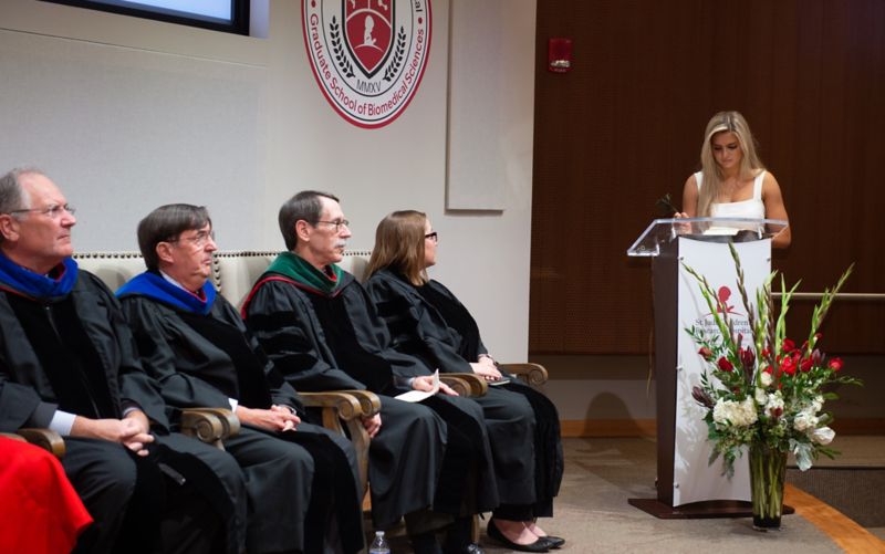 Woman at podium. People in robes sitting to the left