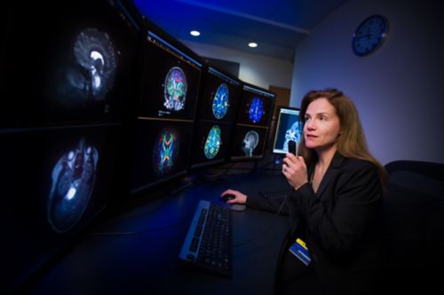 Radiologist sits at desk and reviews brain scans on multiple screens.