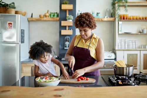 Girl interacting with mother in kitchen as she slices tomatoes on cutting board for salad and boils water for pasta.
