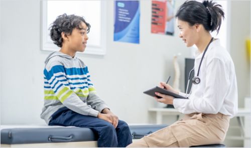 A healthcare professional sitting beside a young patient taking notes on a clipboard.