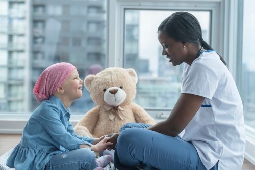 Female child cancer patient interacts with nurse with teddy bear in background