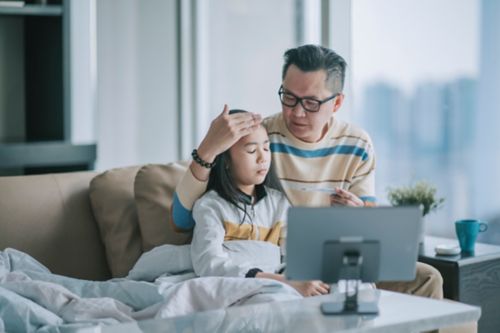 Father taking temperature of daughter on couch while video calling doctor on tablet