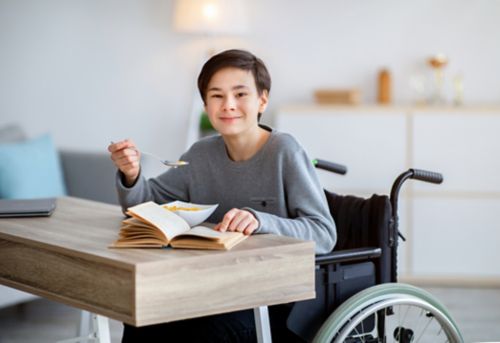 Un adolescente con discapacidad en silla de ruedas comiendo cereales para el desayuno y leyendo un libro en casa.