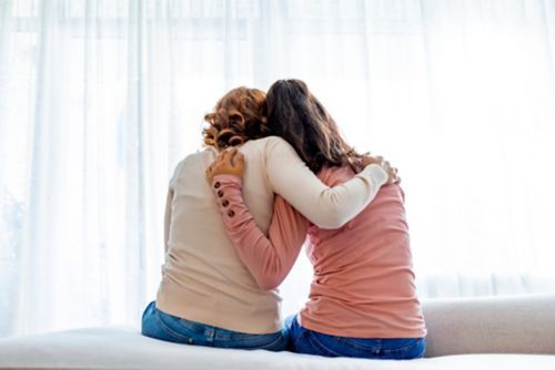 Rear back view of a mother and daughter embrace sitting on bed at home.