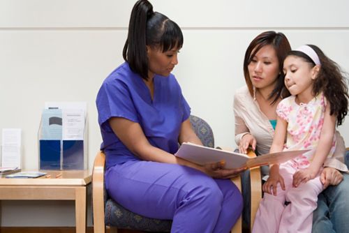 Female nurse in purple scrubs showing a book to a mother and daughter.
