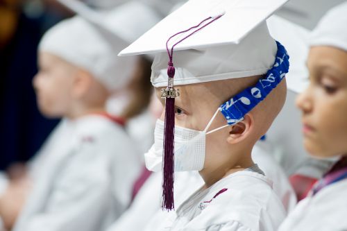 Young pediatric cancer patients sits with classmates in cap, gown, and face mask.