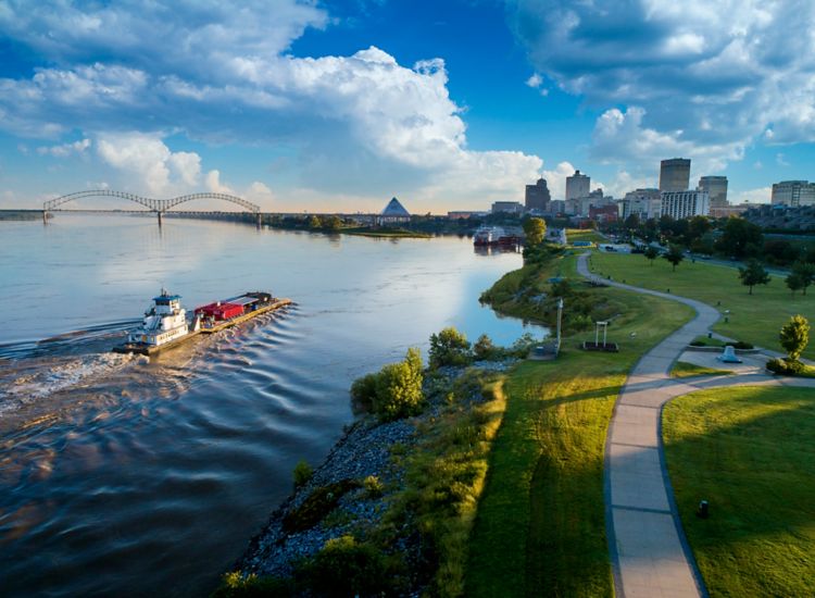 Aerial view of a tug boat on the Mississippi River in Memphis with buildings in the background. To the right is a walkway along the river.