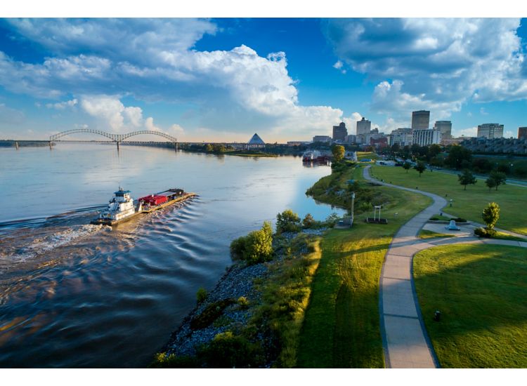 Aerial view of a tug boat on the Mississippi River in Memphis with buildings in the background. To the right is a walkway along the river.