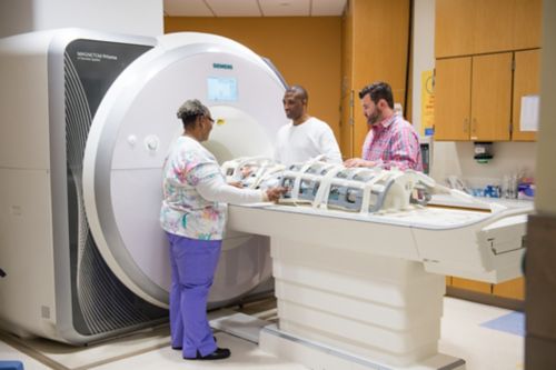 Paciente com câncer infantil posicionado para um exame de RM de corpo inteiro com o pai e dois técnicos de RM ao lado dela.