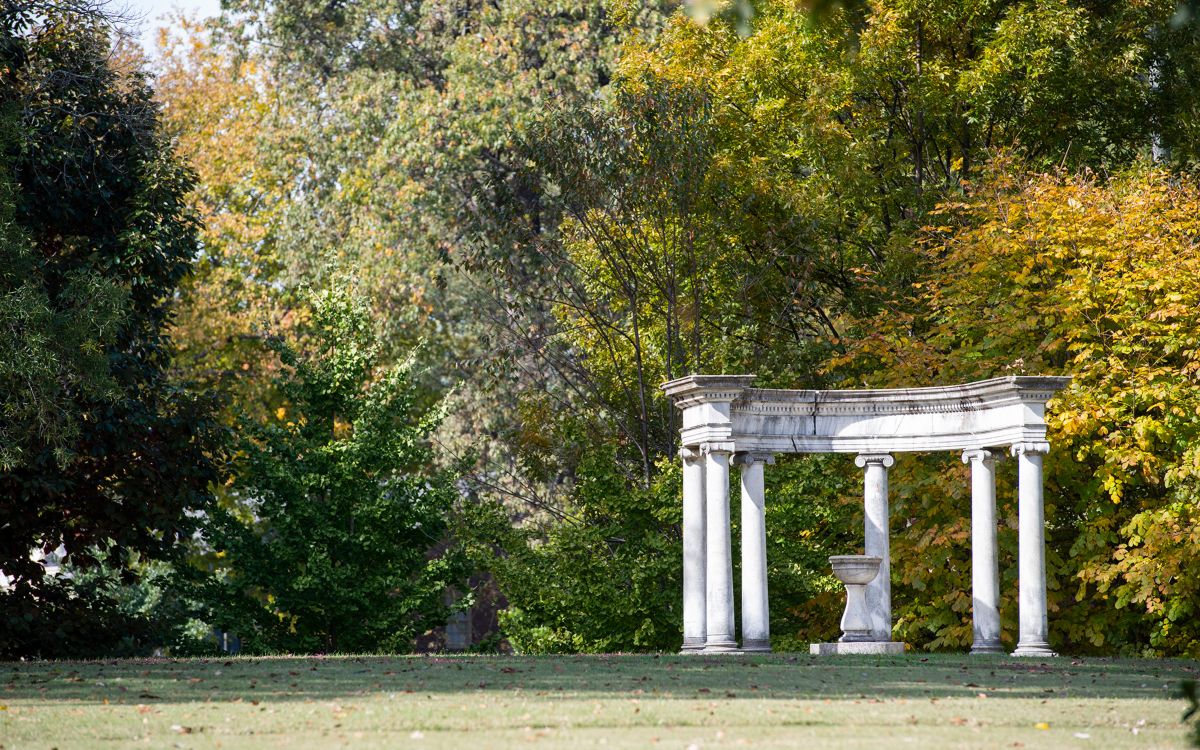 Grecian style columns in a semi circle around a stone urn in the middle of a lawn, surrounded by trees.