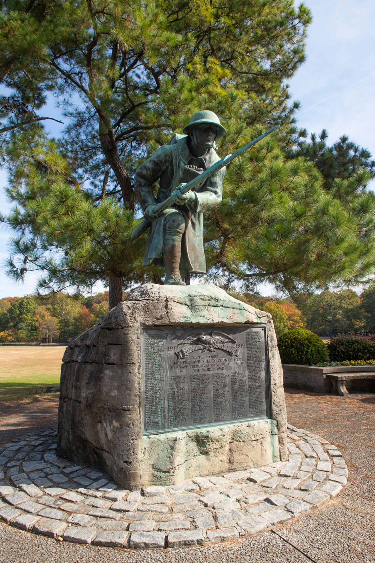 Statue of a solider crouching holding a gun in a plaza, with an pine tree behind.