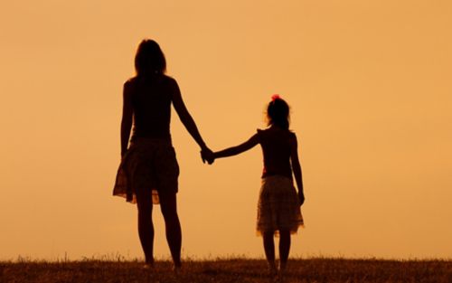 Mother holds hands with young daughter in silhouette.