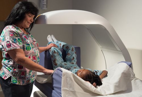 Jovem paciente afro-americano se prepara para passar por uma varredura de densidade óssea com tecnólogo próximos.