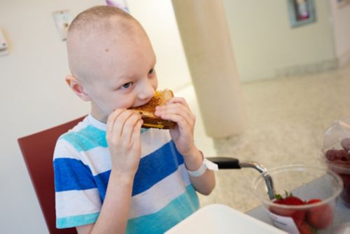 Help children plan eating around times when they are feeling better. Photo shows a childhood cancer patient eating a grilled cheese sandwich in the hospital cafeteria.