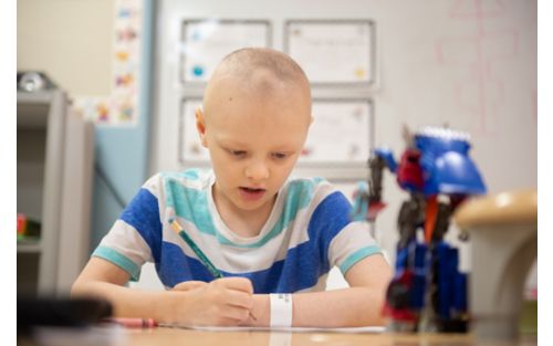 Child cancer patient sitting at school desk with open textbook.